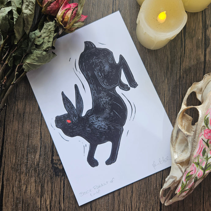 Black rabbit illustration on a white sheet of paper with candles and dried flowers on a wooden surface